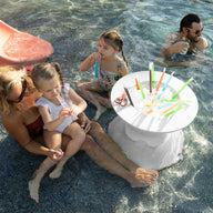 A group of adults and children enjoy a sunny day in a shallow pool. A woman and child sit near a red slide under Ledge Lounger umbrellas. In the water, a Signature Tall Ice Bin Side Table holds drinks with colorful straws. Two men and two children swim nearby.
