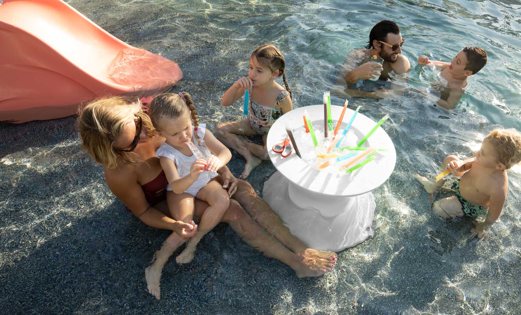 A group of adults and children enjoy a sunny day in a shallow pool. A woman and child sit near a red slide under Ledge Lounger umbrellas. In the water, a Signature Tall Ice Bin Side Table holds drinks with colorful straws. Two men and two children swim nearby.