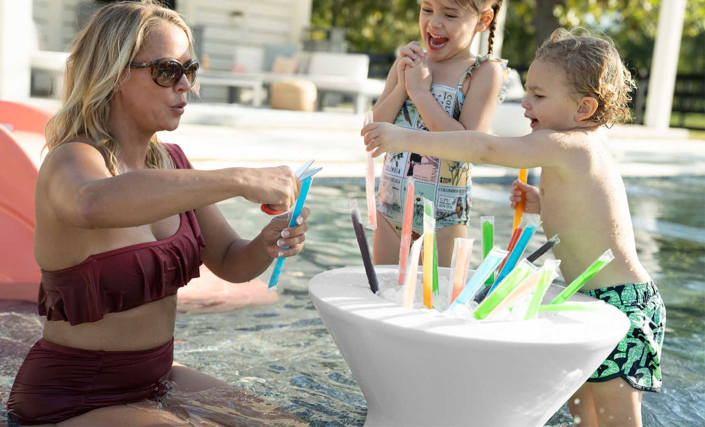 A woman in a maroon swimsuit stands in a pool with two children, selecting colorful ice pops from a Ledge Lounger Signature Tall Ice Bin Side Table. The girl wears a patterned swimsuit and the boy, swim trunks. Ledge Lounger Umbrellas provide shade over the lively patio area in the background.