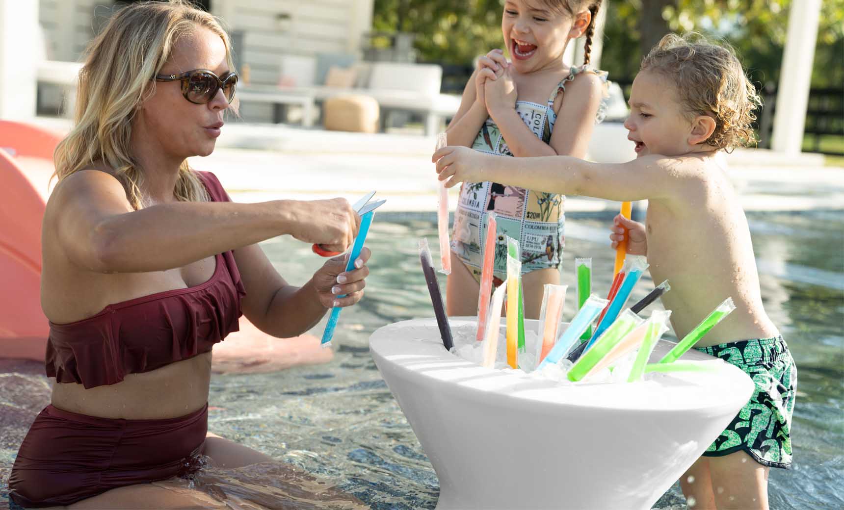 A woman in a maroon swimsuit stands in a pool with two children, selecting colorful ice pops from a Ledge Lounger Signature Tall Ice Bin Side Table. The girl wears a patterned swimsuit and the boy, swim trunks. Ledge Lounger Umbrellas provide shade over the lively patio area in the background.