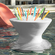 A Ledge Lounger Signature Tall Ice Bin Side Table floats in a pool filled with colorful freezer pops. In the background, a red slide leads into the water, and part of a building is visible.
