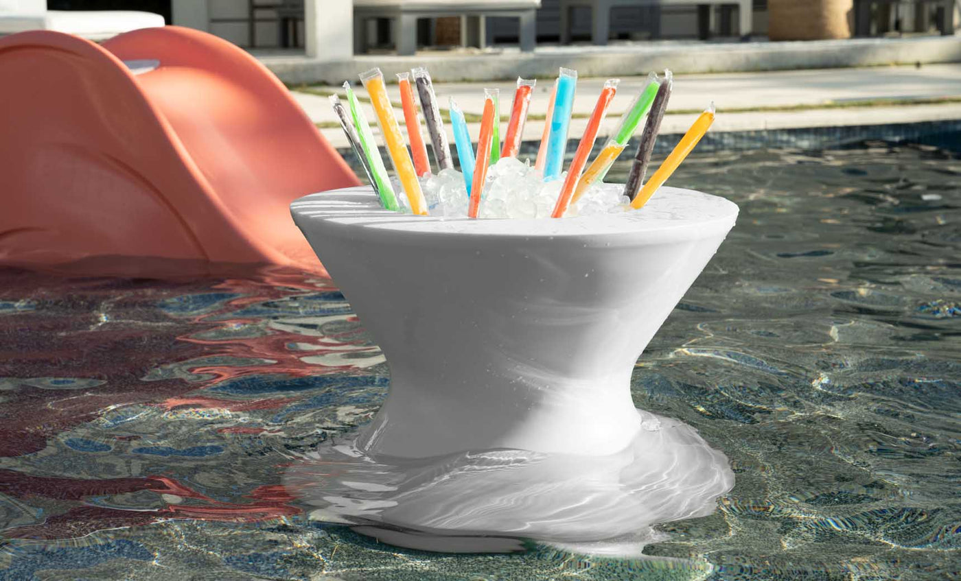 A Ledge Lounger Signature Tall Ice Bin Side Table floats in a pool filled with colorful freezer pops. In the background, a red slide leads into the water, and part of a building is visible.