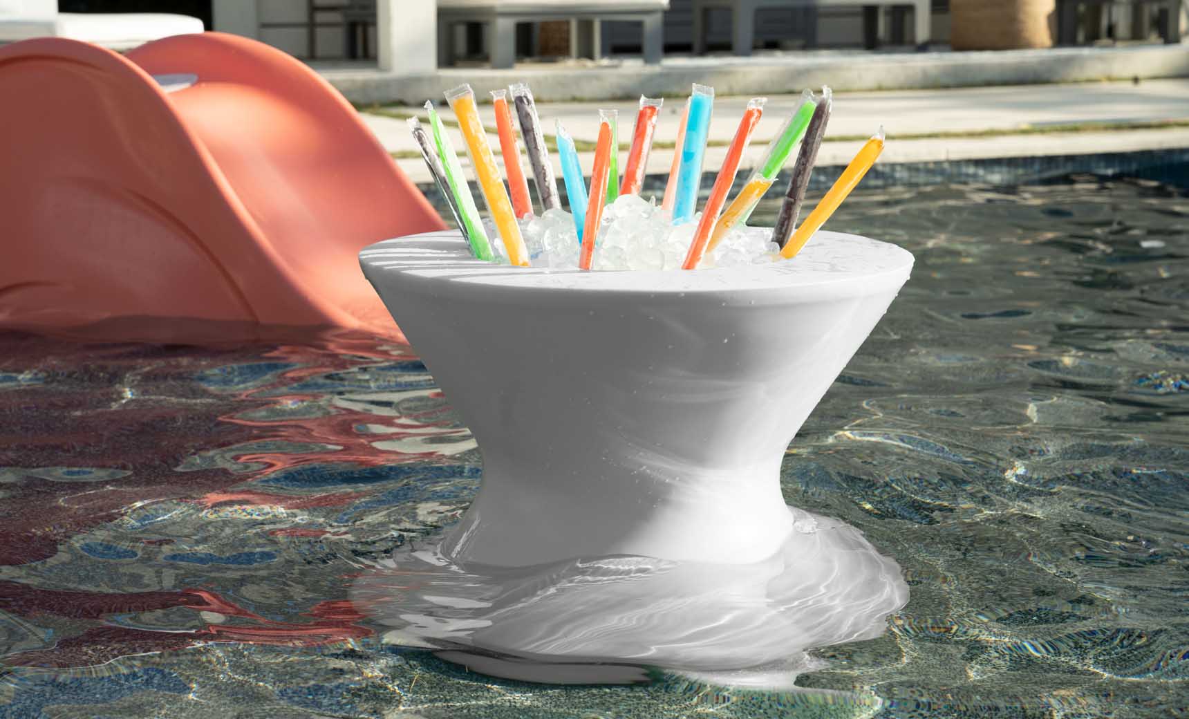 A Ledge Lounger Signature Tall Ice Bin Side Table floats in a pool filled with colorful freezer pops. In the background, a red slide leads into the water, and part of a building is visible.