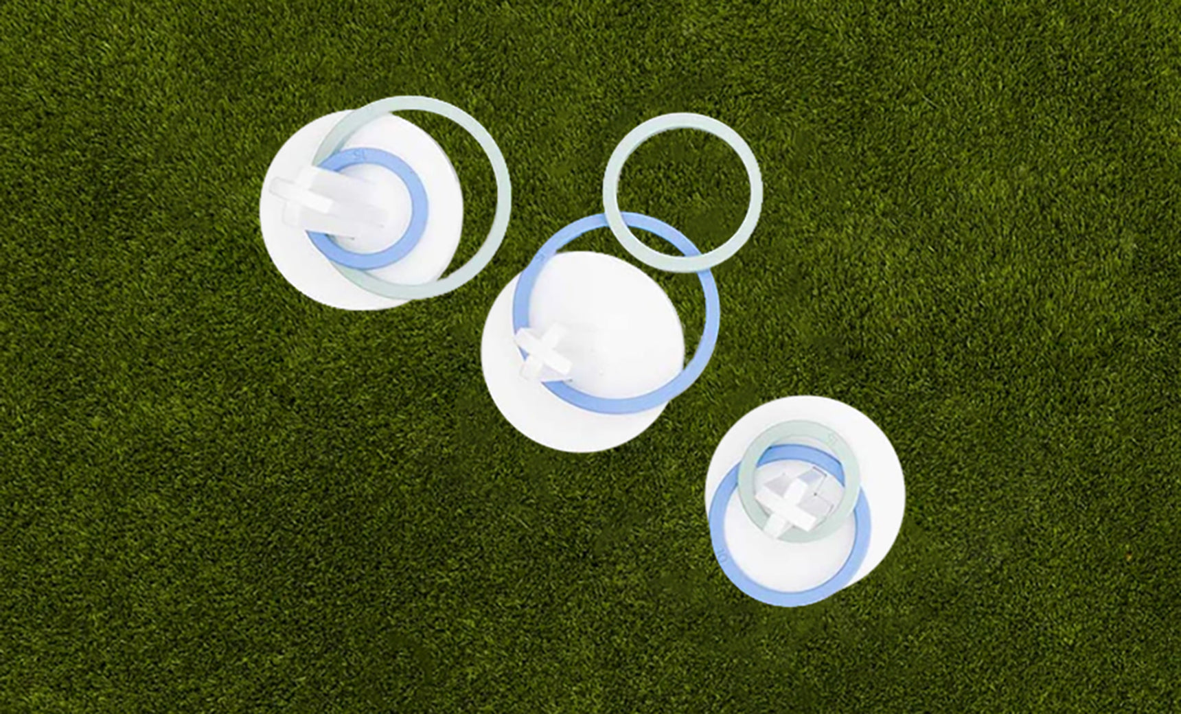 Three white circular discs, featuring blue and white rings and crosses, are arranged on green grass. Their shadow effect adds depth for the ultimate backyard Ring Toss experience, presented by Ledge Lounger.