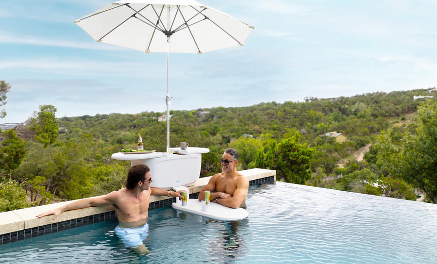 Two men relax in an infinity pool on submerged benches, using the PartyPad Swim-Up Bar by Ledge Lounger as a beverage center with drinks and a large umbrella. The pool offers scenic views of trees and hills under a clear sky.