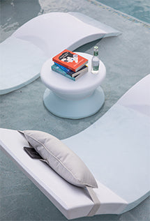 Two white lounge chairs partially submerged in water face each other with a small round table in between. On the table are a stack of books and a bottle of water. A gray cushion rests on one of the chairs.