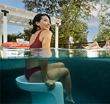 A woman in a red swimsuit sits submerged on a white chair in a clear pool, smiling and looking back. Another person relaxes on a lounge chair in the background. Trees and a white pergola are visible. The scene is sunny and relaxing.