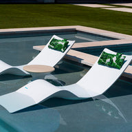 Two modern white lounge chairs, similar to the Ledge Lounger Autograph Chaise, sit partly submerged in a pool with a round table between them. Green leaf-patterned cushions adorn the chairs, and lush grass surrounds this tranquil outdoor setting, enhancing the relaxing atmosphere.