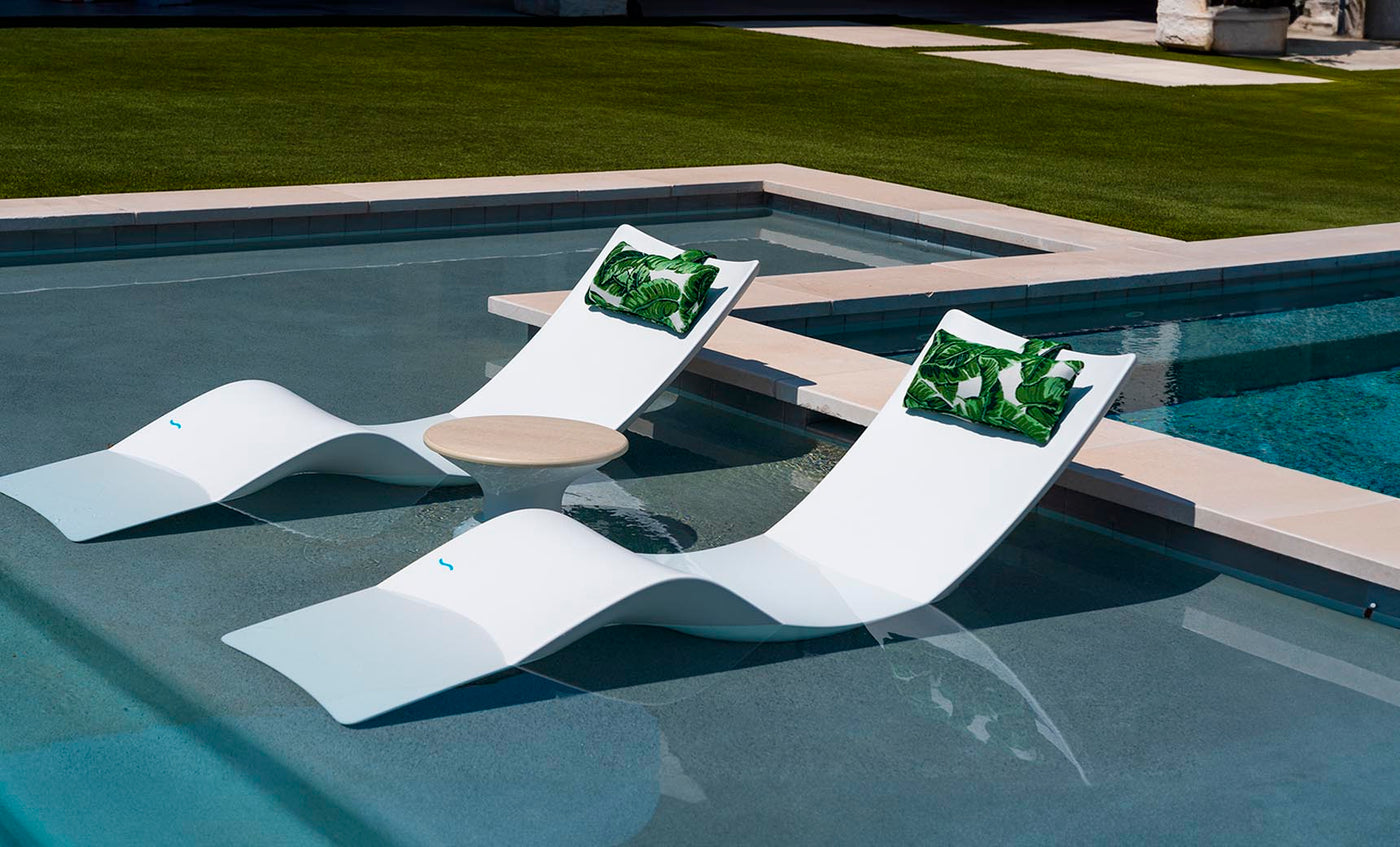 Two modern white lounge chairs, similar to the Ledge Lounger Autograph Chaise, sit partly submerged in a pool with a round table between them. Green leaf-patterned cushions adorn the chairs, and lush grass surrounds this tranquil outdoor setting, enhancing the relaxing atmosphere.