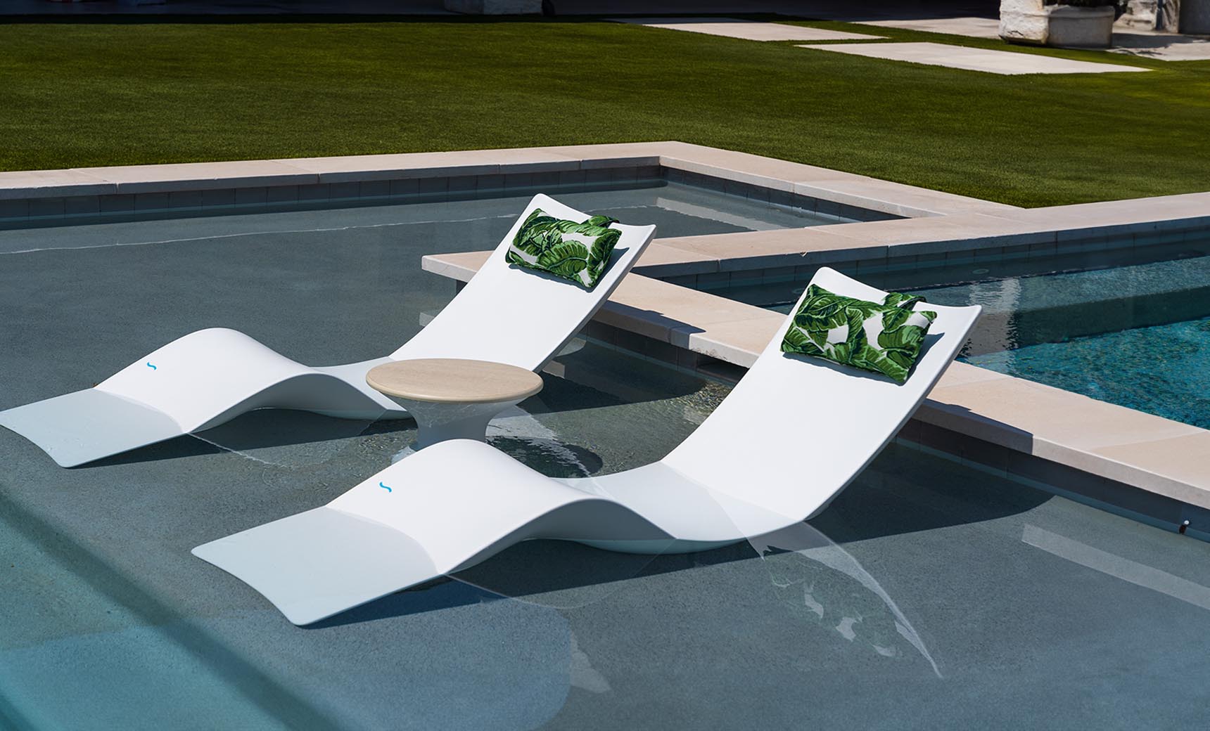 Two modern white lounge chairs, similar to the Ledge Lounger Autograph Chaise, sit partly submerged in a pool with a round table between them. Green leaf-patterned cushions adorn the chairs, and lush grass surrounds this tranquil outdoor setting, enhancing the relaxing atmosphere.