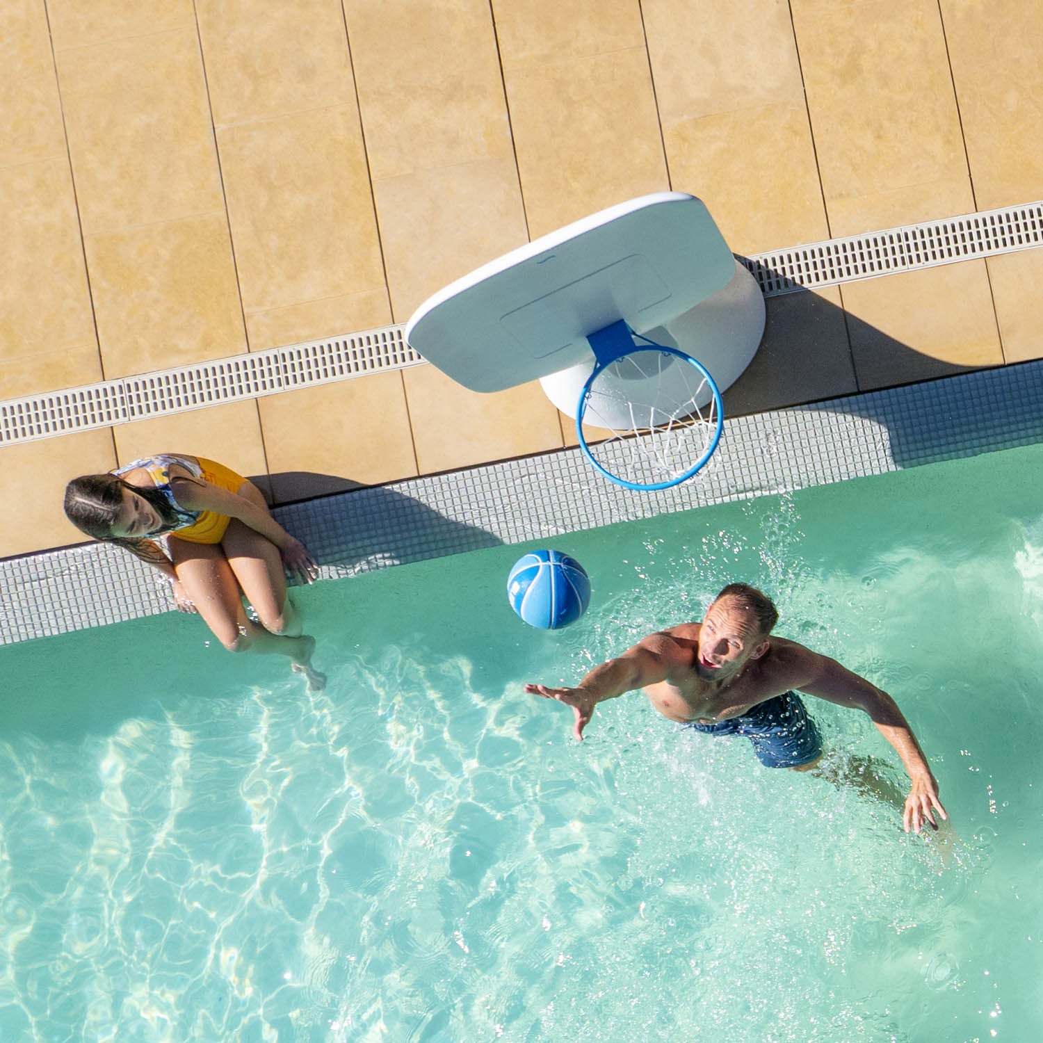 Overhead view of a man in a swimming pool reaching to catch a blue basketball near a floating basketball hoop. A woman sits at the edge of the pool, watching. The pool is surrounded by beige tiles.