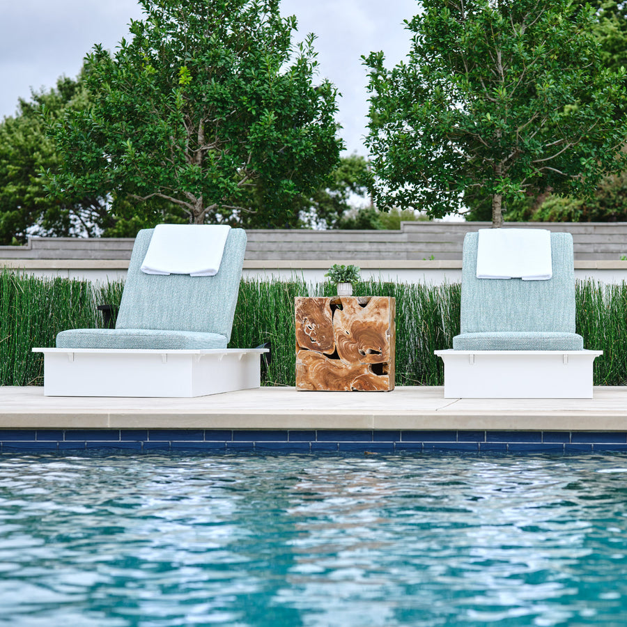 Two cushioned lounge chairs with white towels are positioned by a pool, separated by a square wooden side table. Greenery and trees are visible in the background on a sunny day.