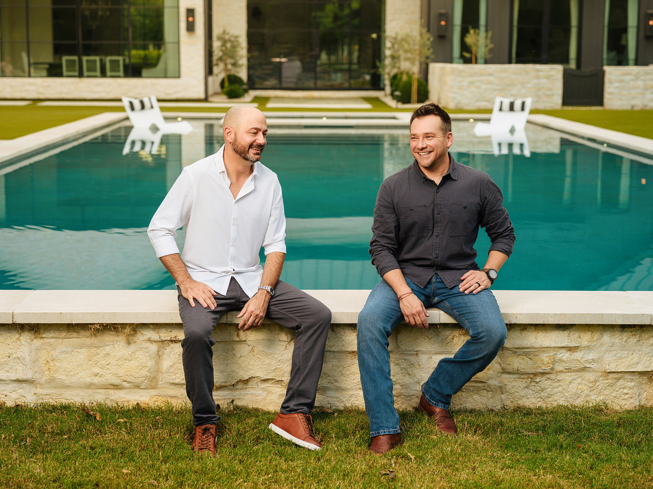 Two men are sitting on the edge of a stone wall in front of a pool. One wears a white shirt and gray pants, and the other wears a black shirt and jeans. Both are smiling, and the setting is outdoors with green grass and a modern building in the background.