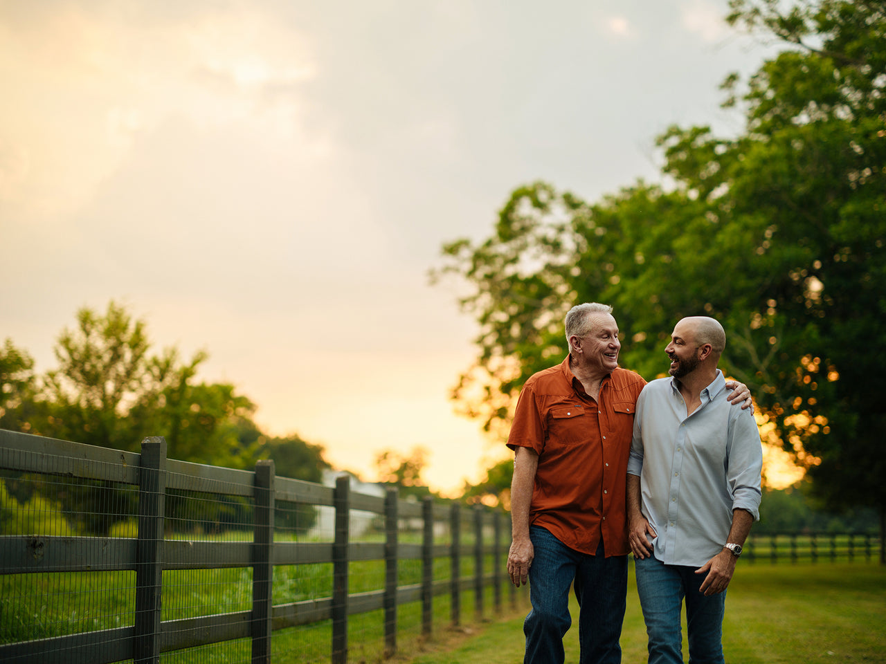 Two men walk along a fenced path during sunset, smiling at each other. One wears an orange shirt, and the other a light blue one. They are surrounded by greenery, with a cloudy sky in the background.