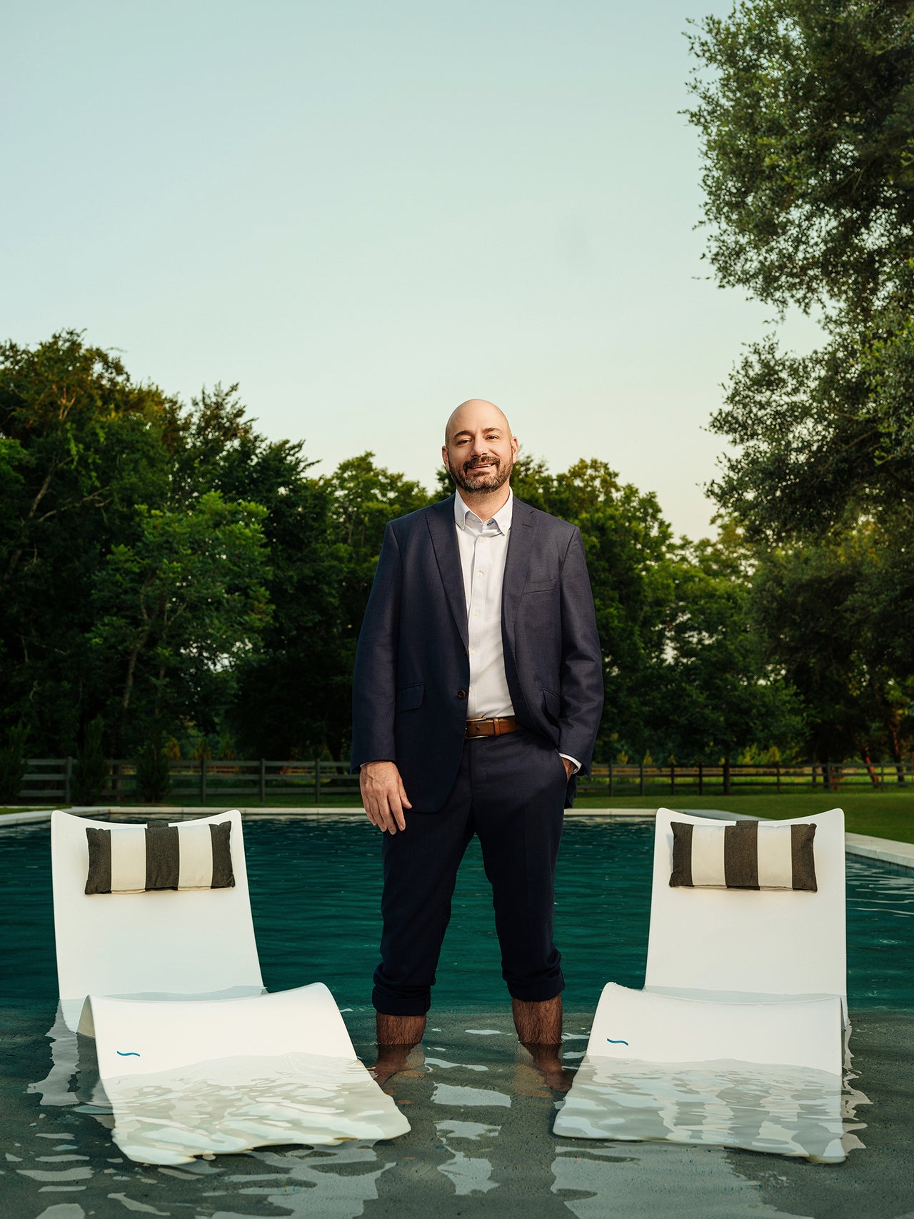 A man in a suit stands ankle-deep in a pool between two white loungers with striped pillows. The background features lush green trees under a clear sky.