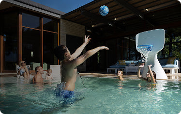 A group of children play basketball in a swimming pool while a few adults watch from the poolside near a modern house with large windows.