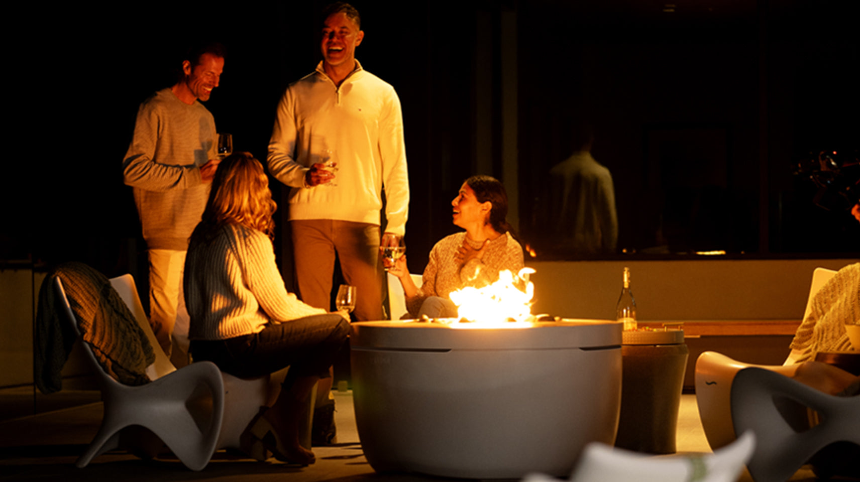 Four people gathered around a lit fire pit at night. Two are standing, holding drinks, and two are seated. They are dressed in casual, warm clothing. The setting is outdoors with modern chairs and a cozy atmosphere.