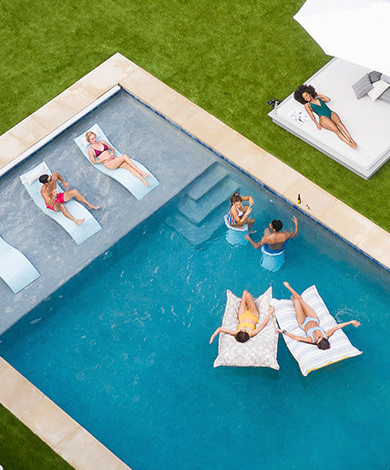 Aerial view of six people relaxing at a pool; four are in the pool on floats or chairs, while two lounge on chairs beside the pool. Another person relaxes on a sofa on the grass nearby.