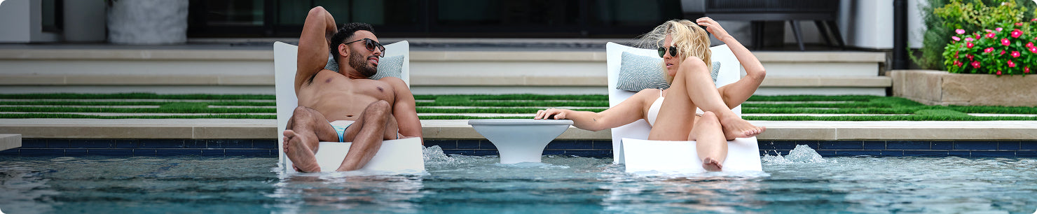 A man and a woman relax on lounge chairs partially submerged in a pool, facing each other and chatting. They both wear sunglasses and swimsuits, with a small table between them. Greenery and flowers are visible in the background.