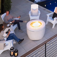 A man and woman relax on the patio in comfortable Adirondack chairs, enjoying wine by a lit fire pit table from the Halo Aglow Bundle by Ledge Lounger. Nearby is a pool, with decorative lights along the fence and lanterns enhancing the ambiance.