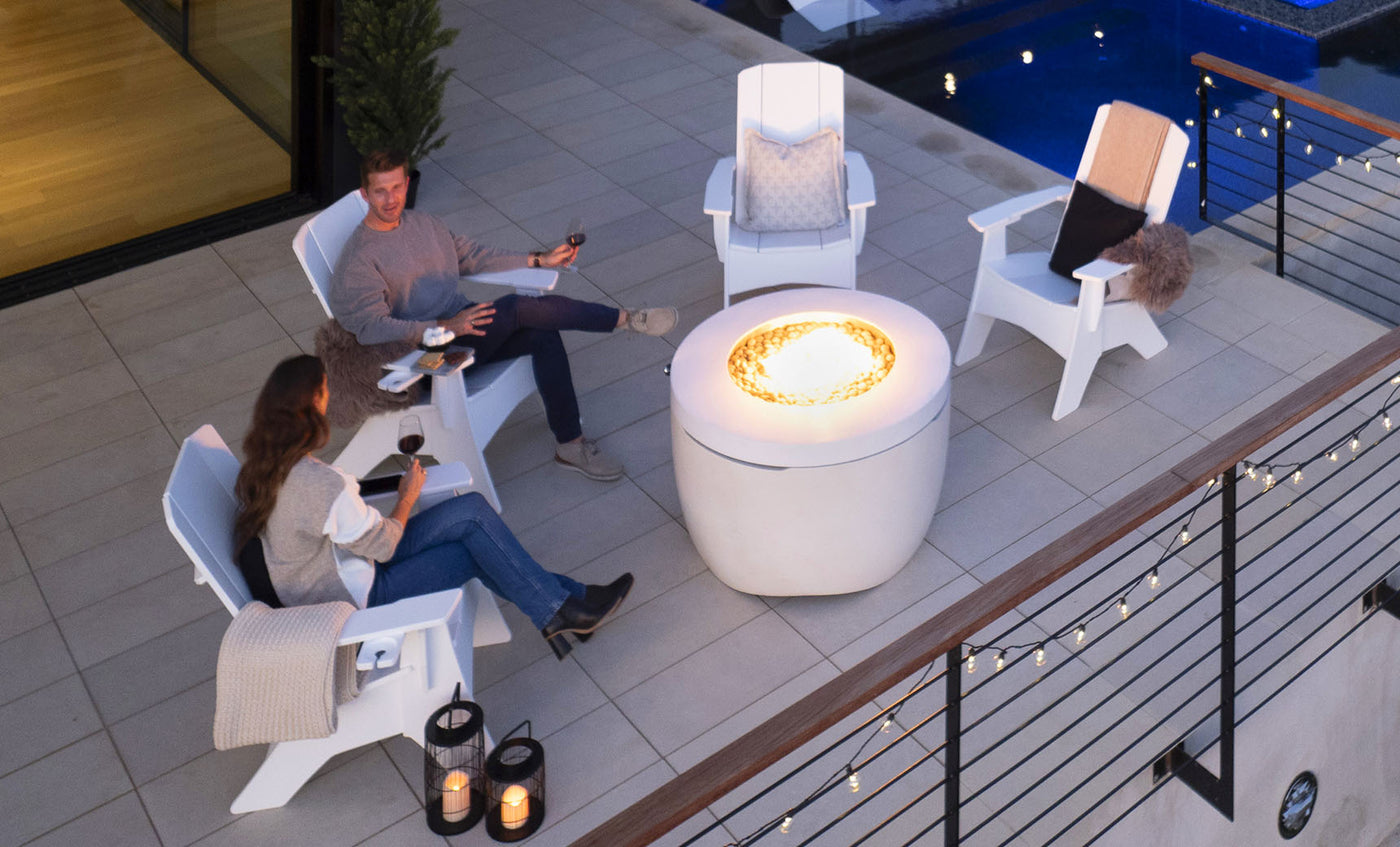A man and woman relax on the patio in comfortable Adirondack chairs, enjoying wine by a lit fire pit table from the Halo Aglow Bundle by Ledge Lounger. Nearby is a pool, with decorative lights along the fence and lanterns enhancing the ambiance.