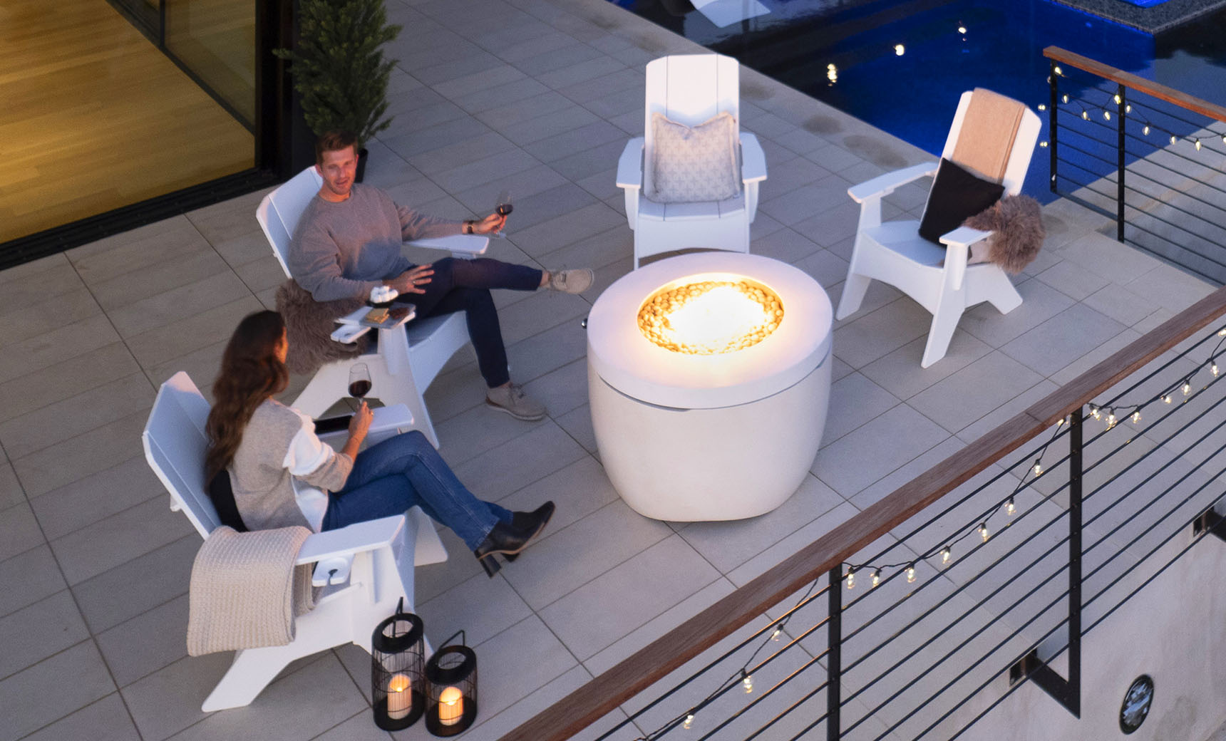A man and woman relax on the patio in comfortable Adirondack chairs, enjoying wine by a lit fire pit table from the Halo Aglow Bundle by Ledge Lounger. Nearby is a pool, with decorative lights along the fence and lanterns enhancing the ambiance.