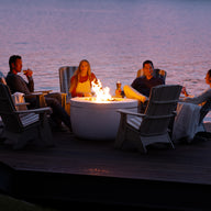 Four friends lounge in Ledge Loungers Halo Aglow Bundle by a calm lakeside at sunset. Seated in Adirondack chairs on the deck, they sip drinks amid a relaxed atmosphere. A large tree stands partially visible on the right.