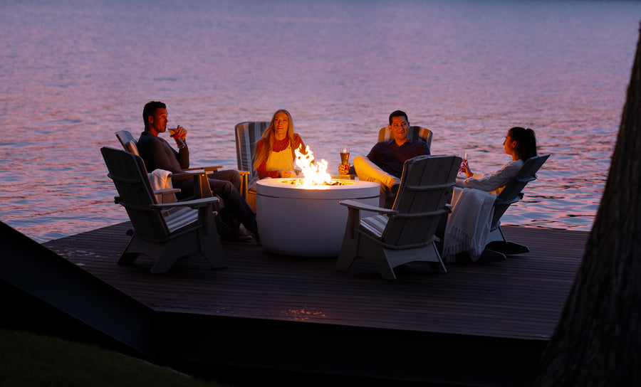 Four friends lounge in Ledge Loungers Halo Aglow Bundle by a calm lakeside at sunset. Seated in Adirondack chairs on the deck, they sip drinks amid a relaxed atmosphere. A large tree stands partially visible on the right.