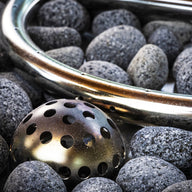 A collection of smooth gray stones encircles a circular metallic object and a dome-like perforated piece, creating an elegant propane fire pit setup. This outdoor ambiance is reminiscent of the Halo Firepit by Ledge Lounger.