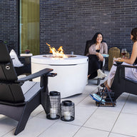 Two people sit in Mainstay Ultimate Adirondack chairs by Ledge Lounger on a modern patio, having a conversation by a circular fire pit. A brick wall and plant create the backdrop, while lanterns and magazines on the ground enhance the charm of this outdoor lounge.