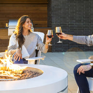 A man and woman sit on Ledge Loungers Halo Firepit in outdoor chairs, clinking glasses of red wine near a barbecue grill. Both are smiling in casual clothes, enhancing the patios warm ambiance.