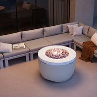 A modern outdoor setup features a gray L-shaped sectional sofa with cushions around Ledge Loungers Halo Firepit, filled with stones. An open book rests on the sofa, while warm lighting and a candle enhance the cozy patio ambiance.