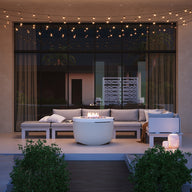 A modern patio with a Ledge Lounger Halo Firepit surrounded by a sectional sofa and illuminated by string lights. A small pool, bordered by potted plants, sits in the foreground, while large windows with sheer curtains cast gentle light on this serene setting.