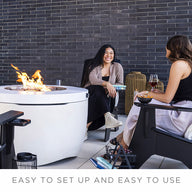 Two people sit on patio chairs with Ledge Loungers round Halo Firepit, enhancing the outdoor ambiance. One holds a glass of wine as they smile and chat against a dark brick wall. Caption: EASY TO SET UP AND EASY TO USE.