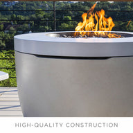 A modern outdoor patio features the sleek Halo Firepit by Ledge Lounger, with a pivot-glide lid and surrounded by a white chair. This serene setup enhances the ambiance, overlooking greenery framed by trees, with HIGH-QUALITY CONSTRUCTION prominently displayed.