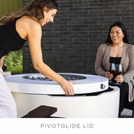 Two women enjoy the outdoors with a Halo Aflame Bundle by Ledge Lounger. One adjusts its lid, while the other sips wine. Theyre surrounded by modern patio furniture and decorative lanterns.