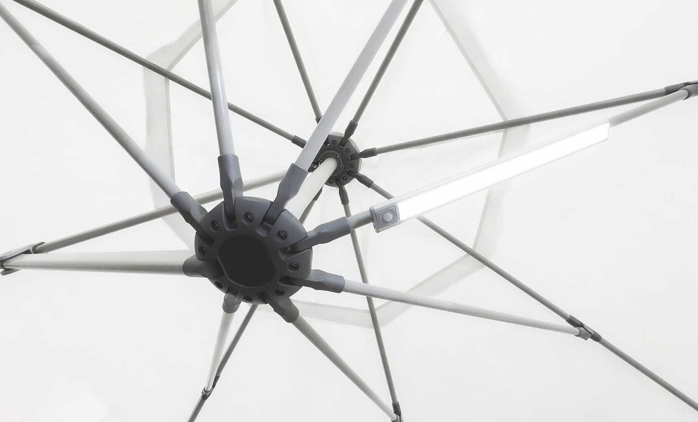 Close-up of the underside of a white umbrella with metal spokes radiating from a central hub. A Ledge Lounger Umbrella Light is attached to one spoke, creating a soft glow against the matching fabric background, ideal for an inviting patio setting.