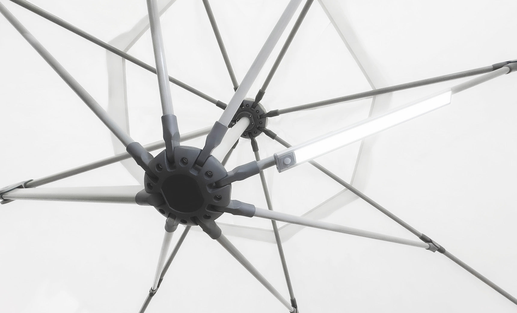 Close-up of the underside of a white umbrella with metal spokes radiating from a central hub. A Ledge Lounger Umbrella Light is attached to one spoke, creating a soft glow against the matching fabric background, ideal for an inviting patio setting.