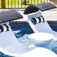 Two white pool lounge chairs are partially submerged in a pool with black canopies and black-and-white pillows. A small Signature Side Table Lid No Hole - Final Sale by Ledge Lounger sits between the chairs. The background features a patio area with a metal fence and grass.