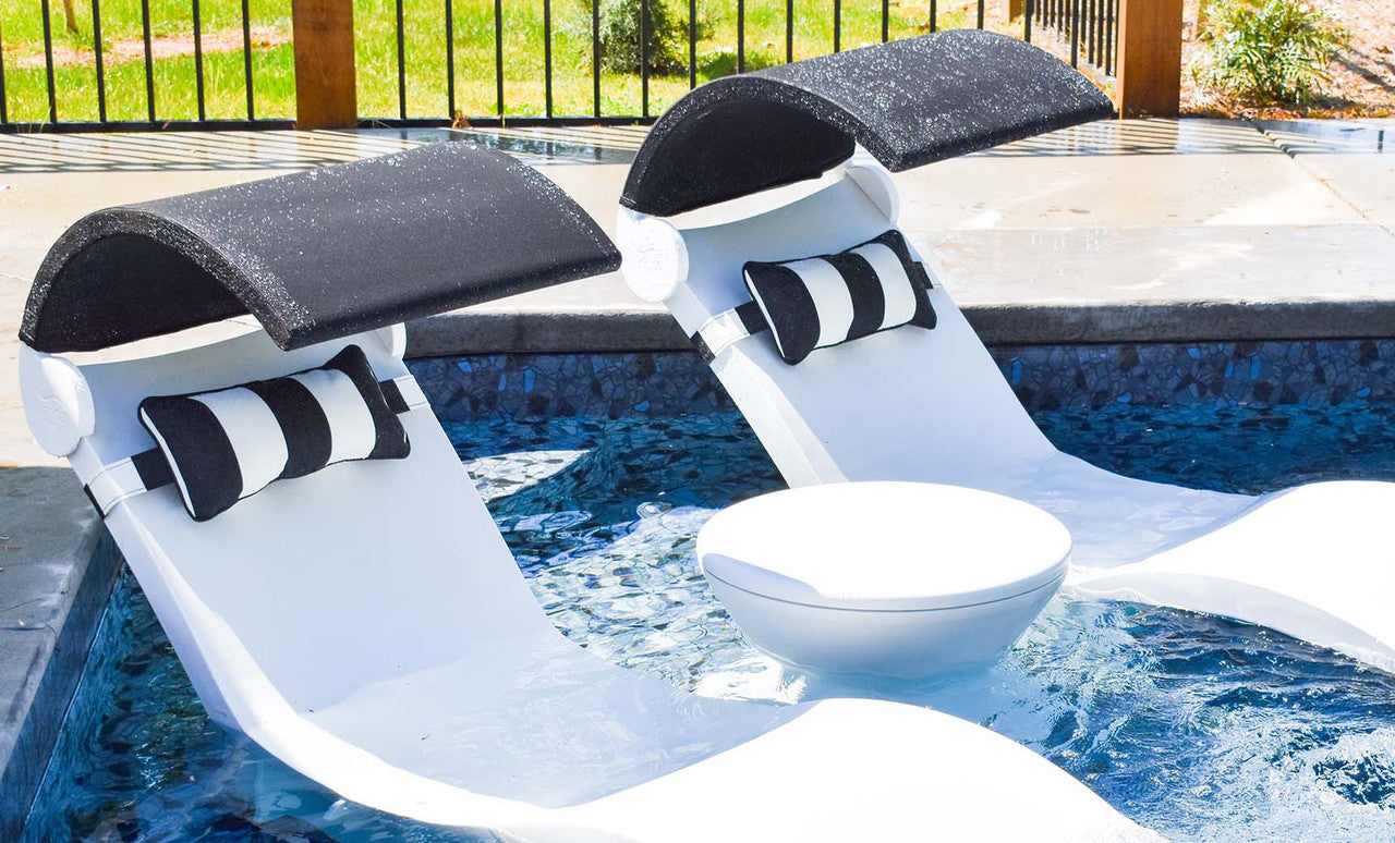 Two white pool lounge chairs are partially submerged in a pool with black canopies and black-and-white pillows. A small Signature Side Table Lid No Hole - Final Sale by Ledge Lounger sits between the chairs. The background features a patio area with a metal fence and grass.