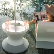 A woman relaxes outside, sitting partially submerged in a pool on a white chair with a wide-brimmed hat, reading a magazine. A round table with the Signature Side Table Lid by Ledge Lounger holds pink drinks under an umbrella reflecting light through its umbrella hole onto the water.