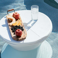 A Signature Side Table Lid with an umbrella hole by Ledge Lounger holds a wooden tray of sliced apples, orange segments, blackberries, and a peach in the pool. A glass of clear water sits beside it. The UV-resistant round white tables shadow adds to the serene and refreshing poolside atmosphere.