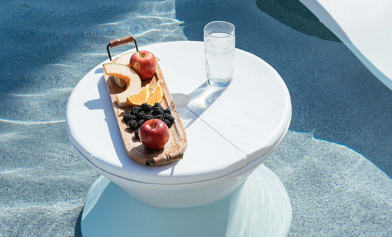 A Signature Side Table Lid with an umbrella hole by Ledge Lounger holds a wooden tray of sliced apples, orange segments, blackberries, and a peach in the pool. A glass of clear water sits beside it. The UV-resistant round white tables shadow adds to the serene and refreshing poolside atmosphere.
