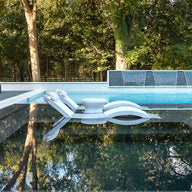 A modern outdoor pool showcases the submerged Signature Chaise from Ledge Lounger, paired with a small round table. Water cascades over a wall, enhancing the serene setting surrounded by lush trees, with additional ergonomic lounge chairs available on a nearby deck for ultimate relaxation.
