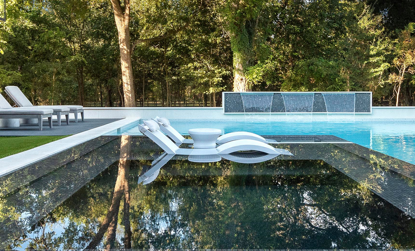 A modern outdoor pool showcases the submerged Signature Chaise from Ledge Lounger, paired with a small round table. Water cascades over a wall, enhancing the serene setting surrounded by lush trees, with additional ergonomic lounge chairs available on a nearby deck for ultimate relaxation.