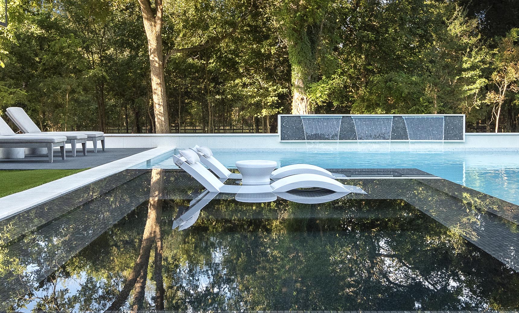 A modern outdoor pool showcases the submerged Signature Chaise from Ledge Lounger, paired with a small round table. Water cascades over a wall, enhancing the serene setting surrounded by lush trees, with additional ergonomic lounge chairs available on a nearby deck for ultimate relaxation.