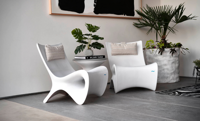 Two modern white chairs with pillows are positioned in a stylish room. A small table between them holds a book and a potted plant. A large planter with green plants is in the background, and a monochrome abstract artwork hangs on the wall.