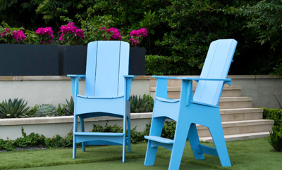 Two blue Adirondack chairs on a grassy area with flowers and plants in the background.
