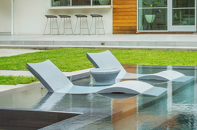 Modern outdoor pool area with submerged gray lounge chairs and a round table. Lawn leads to a wooden-panelled wall and large windows with stools in the background.