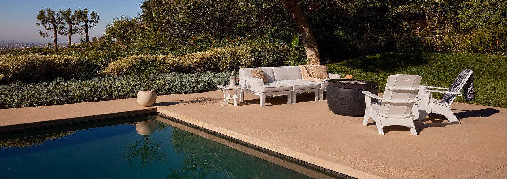 Outdoor patio scene with a swimming pool, white lounge furniture, a dark round coffee table, and a large tree in a landscaped garden under a clear blue sky.